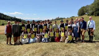 Eine Gruppe von Menschen in traditioneller bayerischer Kleidung, die gelbe Urkunden halten, posiert für ein Foto auf einer Wiese mit malerischer Kulisse.