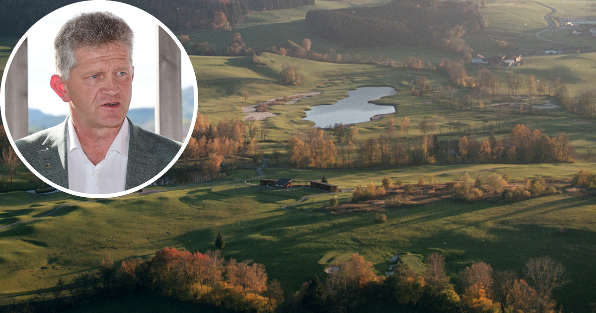Ein Mann in einem Anzug steht vor einem Golfplatz mit einem See und Bäumen im Hintergrund.