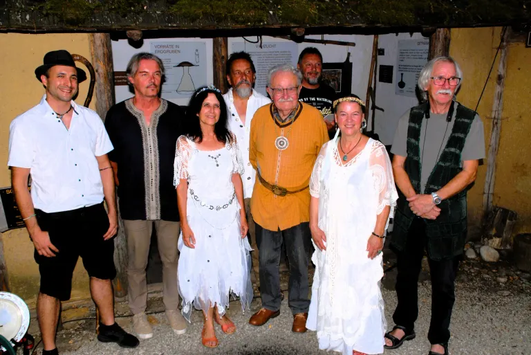 Eine Gruppe von Menschen, die verschiedene Kostüme tragen, darunter weiße Kleider und mittelalterliche Kleidung, posiert für ein Foto vor einem rustikalen Gebäude.