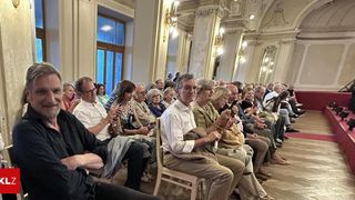 Eine Gruppe von Menschen sitzt in einem Saal, einige mit Brillen und halten Mikrofone. Sie scheinen an einer Diskussion oder Präsentation teilzunehmen. Der Saal ist gut beleuchtet mit Kronleuchtern und hat einen Holzboden.