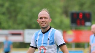 Ein Mann in einem blau-weißen Fußballtrikot mit Logo und Text auf der Brust lächelt und schaut nach vorne.