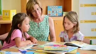 Eine Frau mit Brille unterrichtet zwei junge Mädchen, wie man liest, mit Büchern und bunten Kreisen auf einem Tisch.