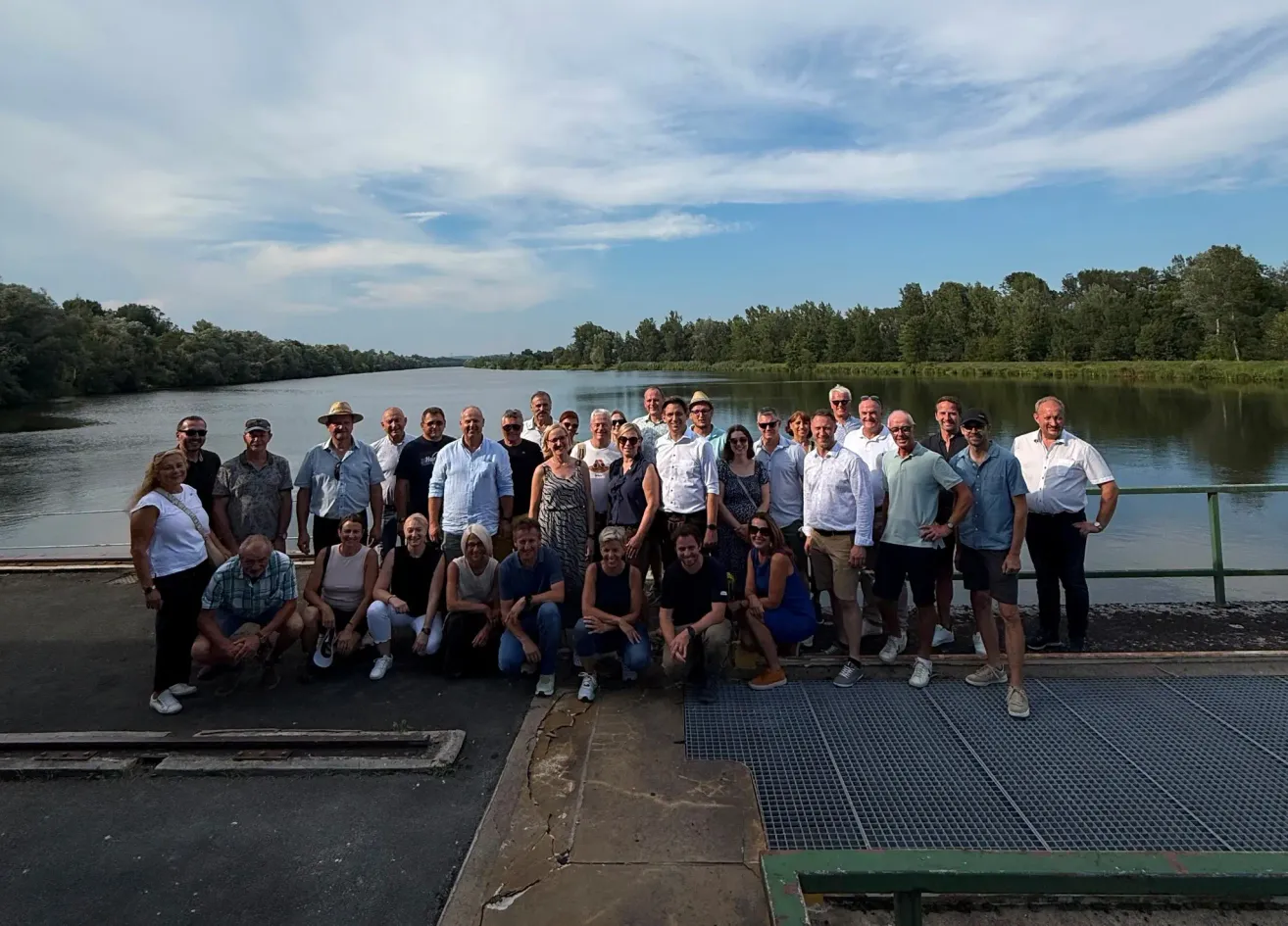 Eine Gruppe von Menschen posiert für ein Foto auf einer Betonplattform in der Nähe eines Flusses mit einer Kulisse aus Bäumen und einem bewölkten Himmel.
