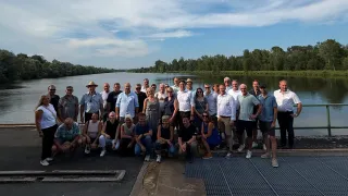 Eine Gruppe von Menschen posiert für ein Foto auf einer Betonplattform in der Nähe eines Flusses mit einer Kulisse aus Bäumen und einem bewölkten Himmel.