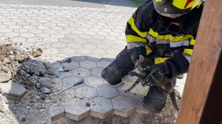 Ein Feuerwehrmann in voller Ausrüstung kniet auf einem rissigen Bürgersteig und hält eine kleine Katze in den Armen. Ein blauer Schraubendreher liegt in der Nähe auf dem Boden.