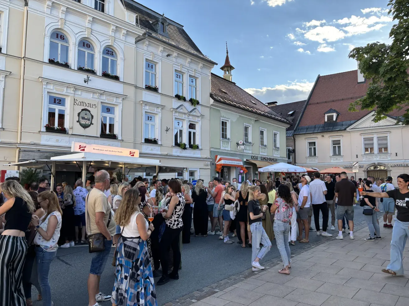 Eine Menschenmenge steht auf einer Straße vor Gebäuden mit Nummern und Schildern, unter einem klaren blauen Himmel. Einige Leute halten Getränke.