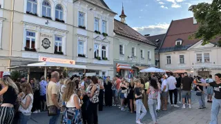 Eine Menschenmenge steht auf einer Straße vor Gebäuden mit Nummern und Schildern, unter einem klaren blauen Himmel. Einige Leute halten Getränke.