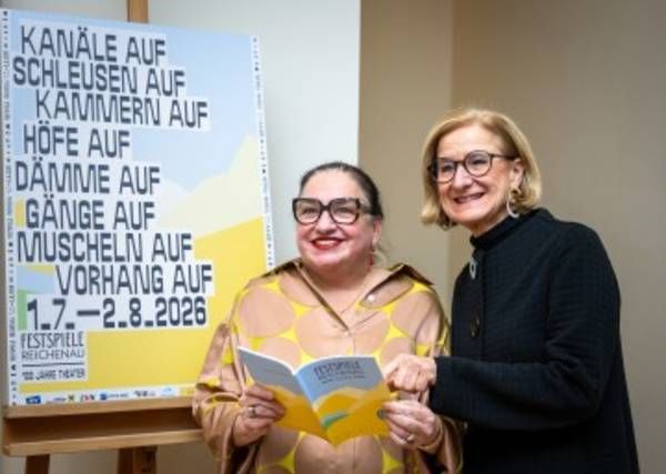 Zwei Frauen stehen neben einem Plakat, eine hält ein Buch. Das Plakat hat deutschen Text und einen Datumsbereich von 1.7-2.8.2026. Die Frau auf der linken Seite trägt ein gelbes Kleid und eine Brille. Die Frau auf der rechten Seite trägt einen schwarzen Mantel und eine Brille.