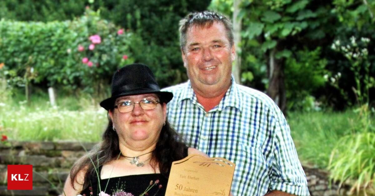 Ein Mann und eine Frau stehen lächelnd nebeneinander, mit einem Zertifikat in der Hand der Frau. Der Mann trägt ein kariertes Hemd, und die Frau trägt einen schwarzen Hut und eine Brille. Hinter ihnen sind grüne Büsche und rosa Blumen.