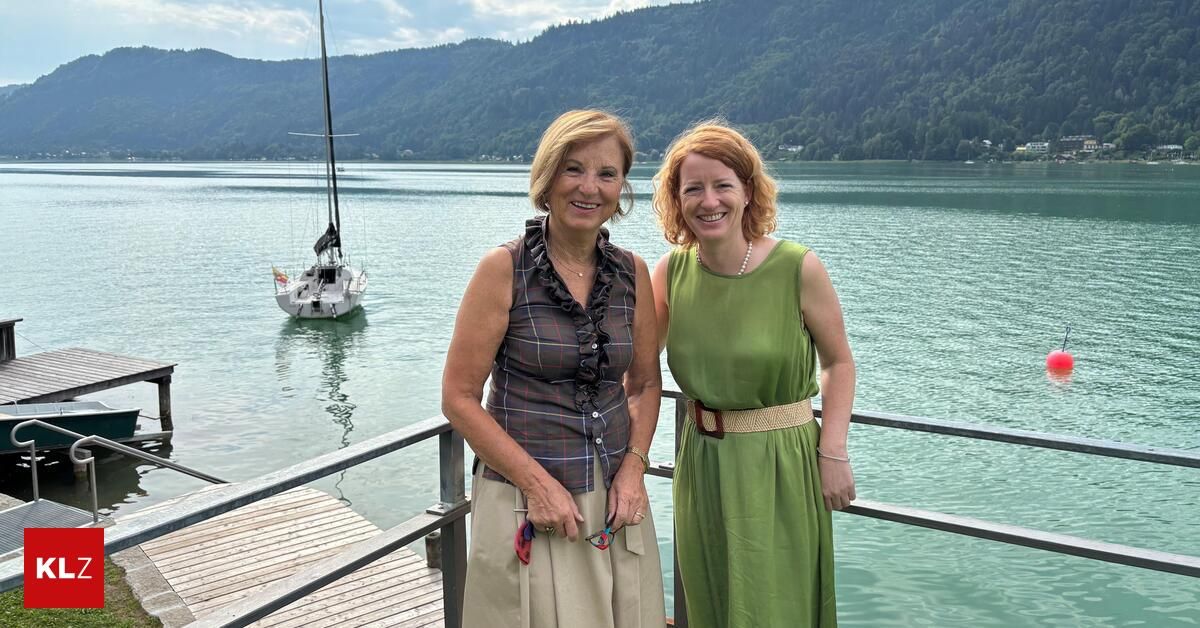 Zwei Frauen stehen auf einem Steg am See mit einem Segelboot im Hintergrund. Die Frau links trägt eine karierte Bluse und einen Gürtel, während die Frau rechts ein grünes Kleid trägt.