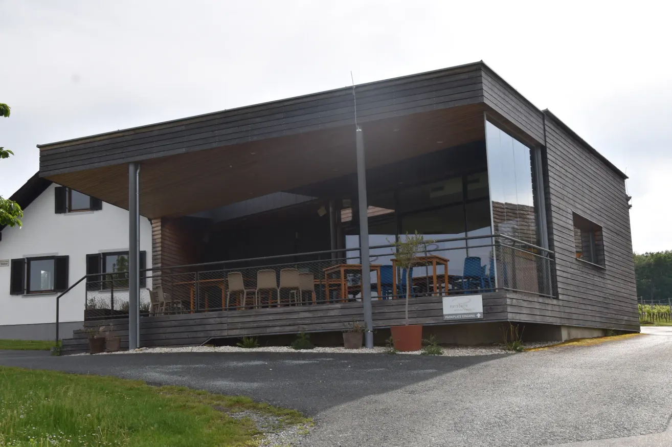 A modern building with a wooden exterior and glass windows, featuring a patio with tables and chairs. The building has a sign with text and a potted plant.
