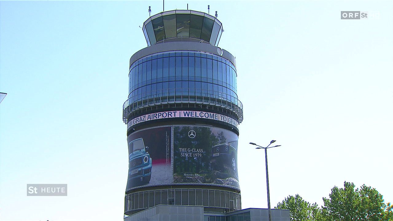 Ein hoher zylindrischer Turm mit einer Mercedes-Benz-Werbung darauf. Der Turm hat Glasfenster und einen Kontrollraum oben. Eine Straßenlaterne befindet sich auf der rechten Seite.