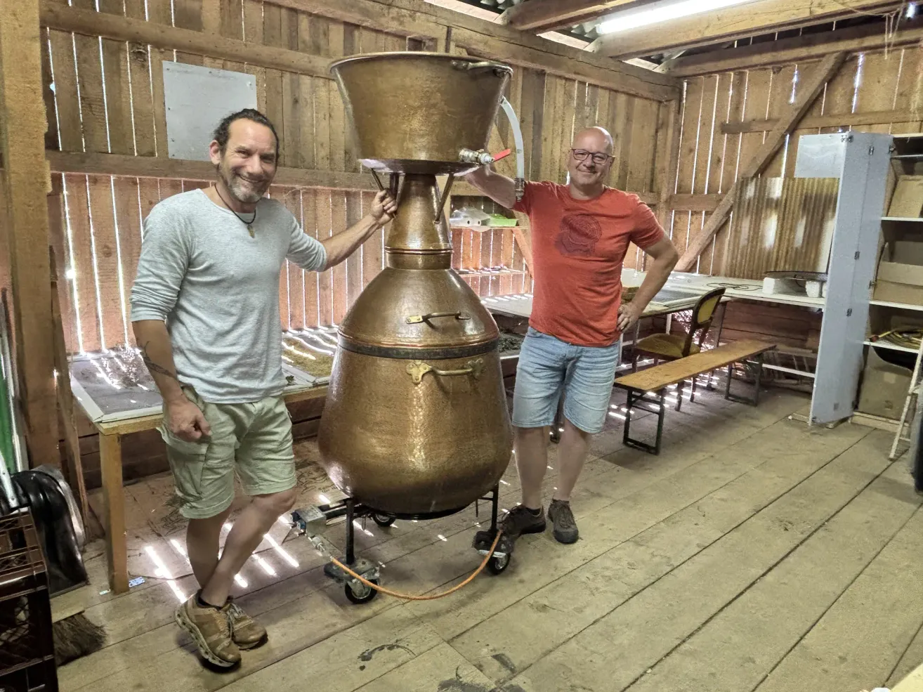 Two men stand beside a large copper pot in a rustic workshop, one smiling, the other with glasses. Behind them, a wooden bench and table are visible.