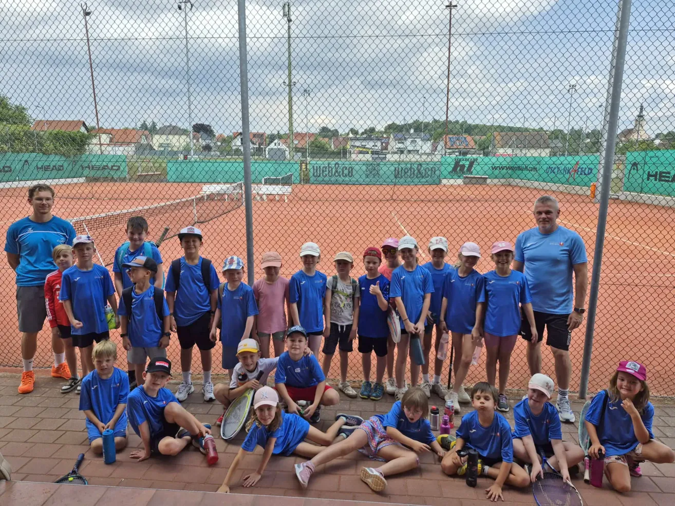 Eine Gruppe junger Tennisspieler in blauen Uniformen posiert für ein Foto auf einem Tennisplatz mit einem Zaun und einem bewölkten Himmel im Hintergrund.
