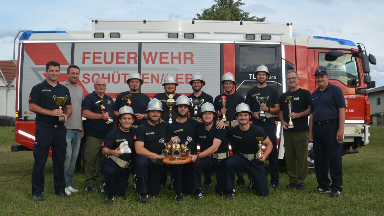 A group of firefighters in uniform, wearing helmets, are posing for a photo with their trophies in front of a fire truck.