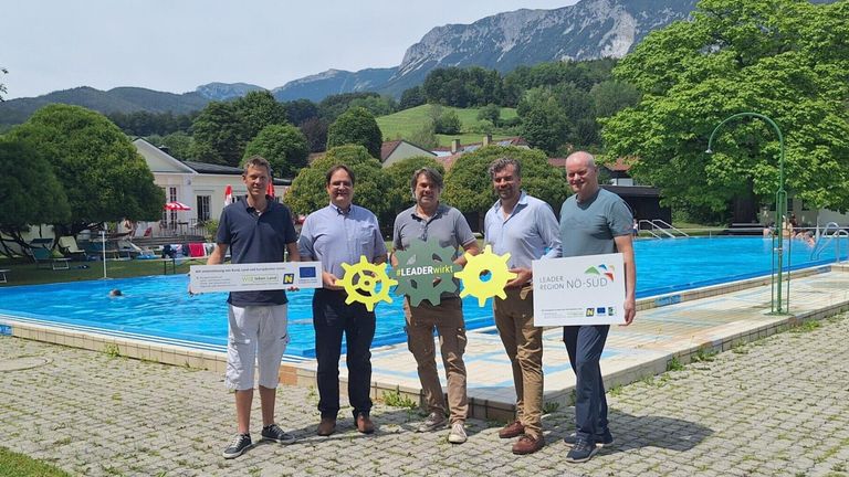 Fünf Männer stehen vor einem Pool und halten Banner mit den Texten LEADER Region NO-SUD und #LEADERwirkt. Sie lächeln und scheinen für ein Foto zu posieren. Dahinter befindet sich eine Bergkette und ein Gebäude.