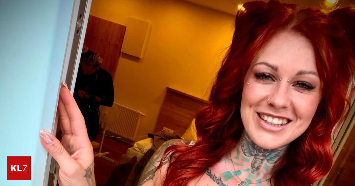 Eine Frau mit roten Haaren und Tattoos am Hals und an den Armen lächelt und posiert für ein Foto. Hinter ihr steht eine Person in der Nähe eines Heizkörpers.