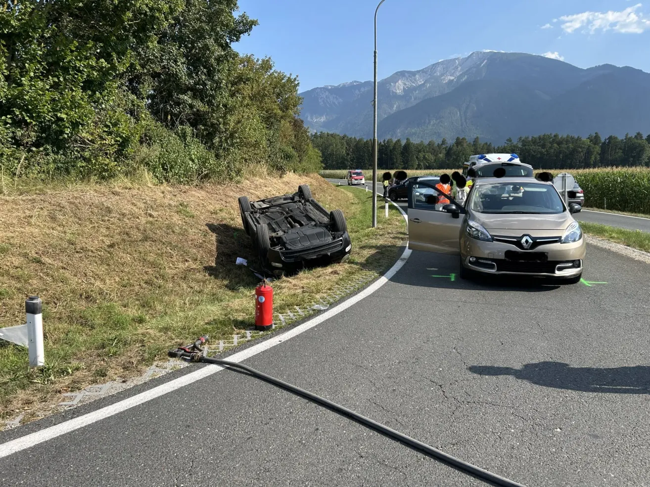 A flipped car rests on the side of a road, with emergency vehicles nearby. A fire extinguisher is placed on the grass.