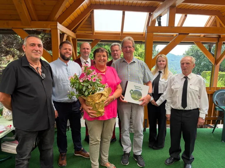 A group of people stand under a wooden gazebo, smiling for a photo. A woman in pink holds a bouquet and a certificate. Behind her, a man in a checkered shirt holds a certificate.