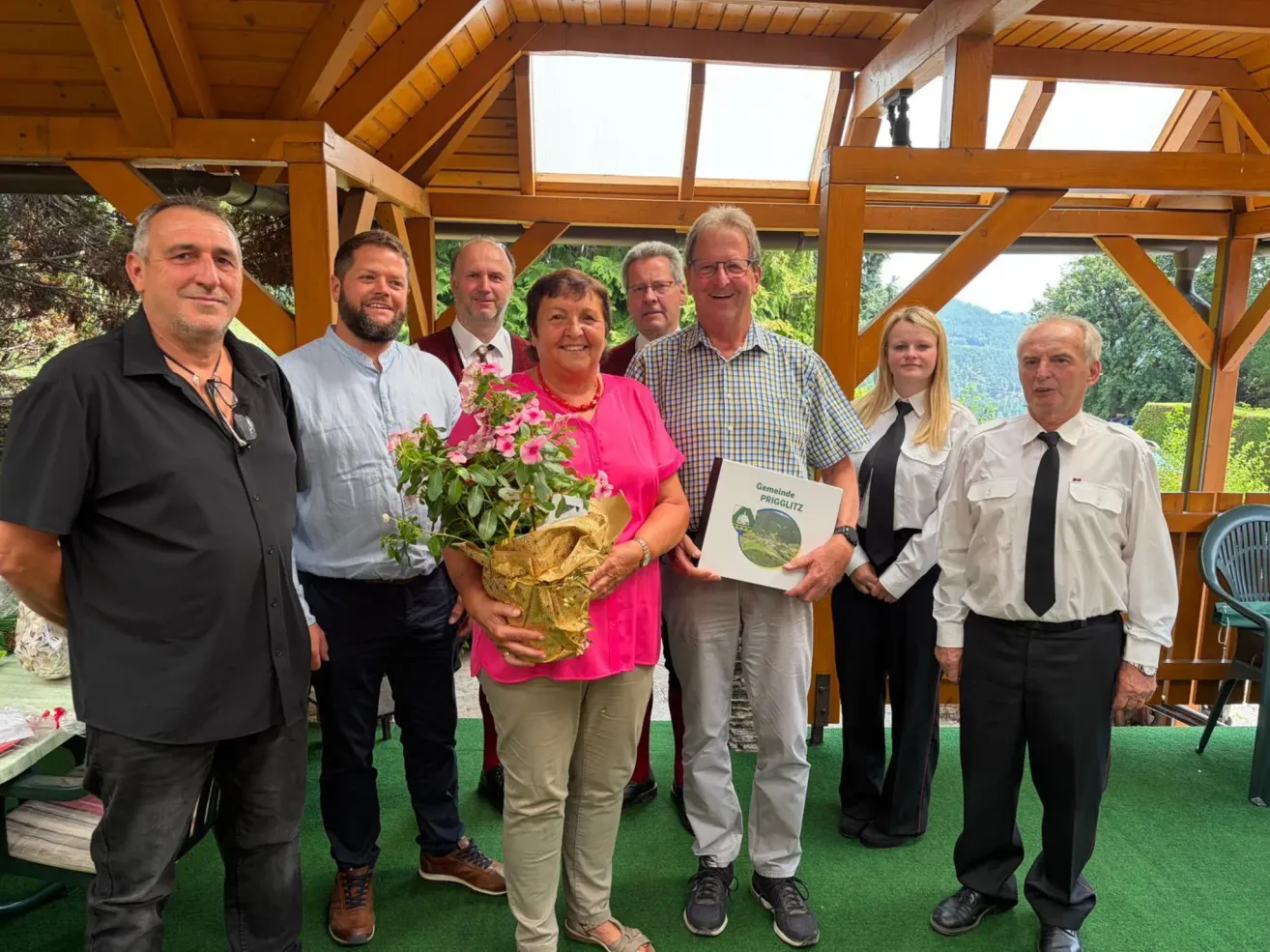 A group of people stand under a wooden gazebo, smiling for a photo. A woman in pink holds a bouquet and a certificate. Behind her, a man in a checkered shirt holds a certificate.