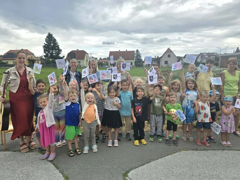 Eine Gruppe von Kindern und Erwachsenen steht in einer Reihe und hält bunte Zeichnungen hoch. Sie sind draußen in der Nähe eines Grasfeldes und Häusern.