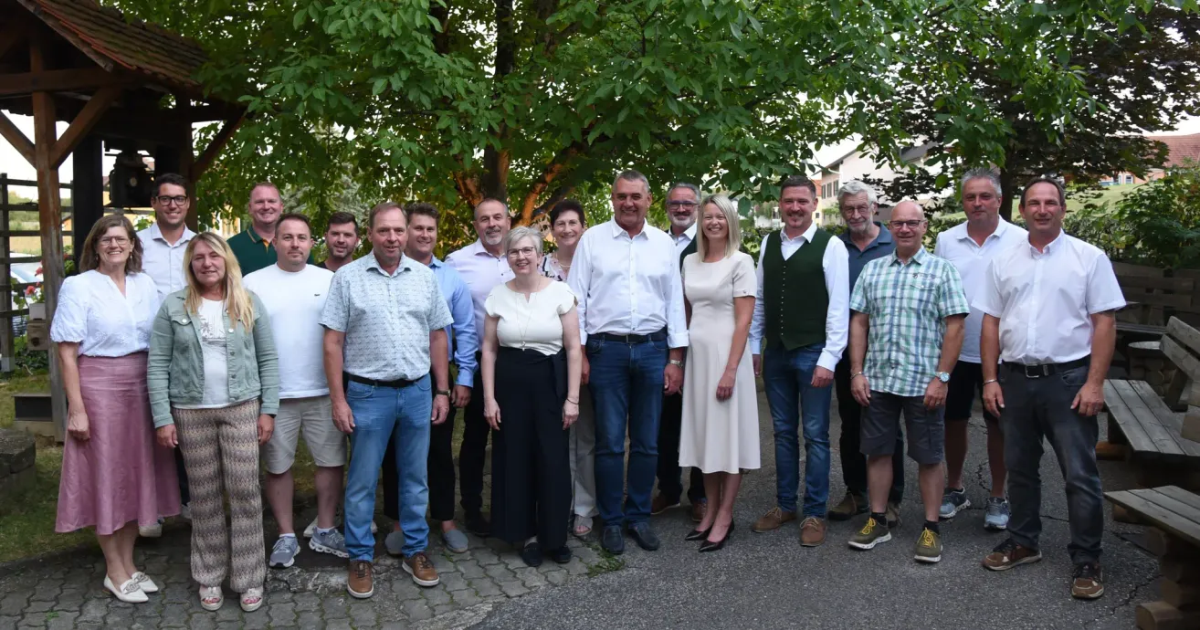 A group of people, including men and women, are standing together under a tree, smiling for a photograph. They are dressed in casual and formal attire.