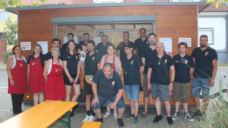 A group of people wearing matching black shirts with logos stand in front of a wooden building. Some wear aprons and glasses, and a few hold a baby. A yellow table and bench are in the foreground.