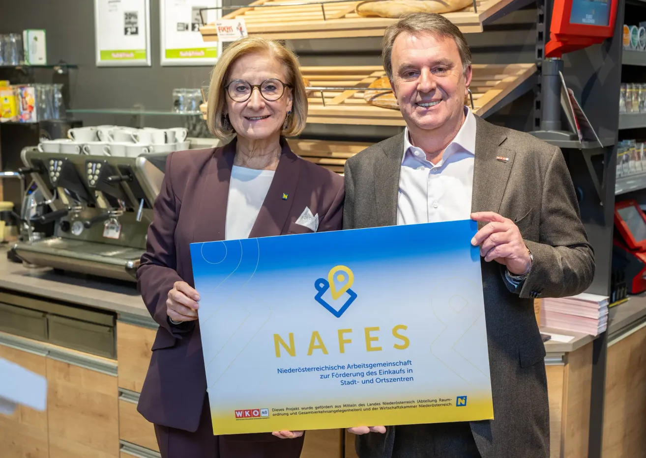 A man and a woman stand in front of a bakery, holding a blue sign that says 'NAFES'. They are smiling and appear to be posing for a photo. Behind them, there are shelves with bread and a coffee machine.