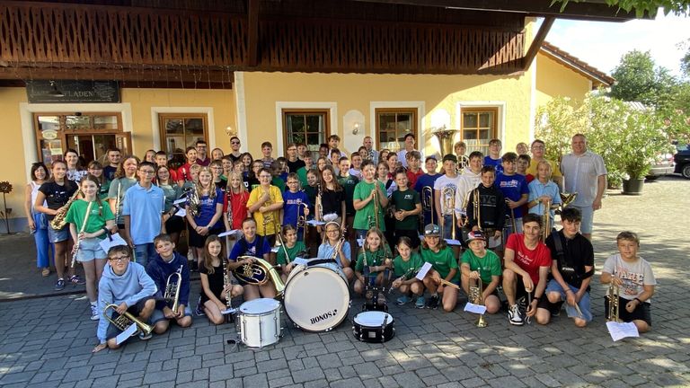 A large group of children and young adults, dressed in green shirts and holding musical instruments, pose for a photo in front of a yellow building.