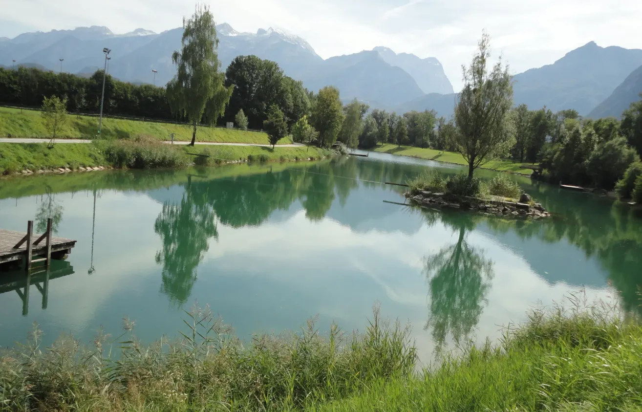 Ein ruhiger See mit klarem Wasser spiegelt die umliegenden Bäume und Berge unter einem teilweise bewölkten Himmel wider.