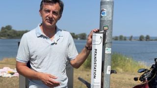 Ein Mann in einem hellblauen Poloshirt steht neben einem Metallpfosten mit einem Schild, das 'Pendstation' besagt. Der Pfosten hat verschiedene Aufkleber und befindet sich in der Nähe eines Gewässers.