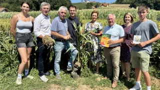 Eine Gruppe von Menschen steht in einem Maisfeld, einige halten Maiskolben und Bücher, mit einem klaren blauen Himmel und Bäumen im Hintergrund.