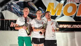 Drei Personen stehen auf einem Podium, lächeln und halten Flaschen Wein in den Händen. Im Hintergrund befindet sich ein Banner mit dem Text 'SPEED MO' und einem Auto-Grafik. Der Mann links trägt grüne Shorts, die anderen tragen schwarze Shorts.