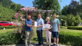 Vier Menschen stehen in einem Garten, lächeln und halten Urkunden. Sie stehen vor einem hölzernen Bogen mit rosa Blumen. Der Mann auf der linken Seite trägt ein blaues Hemd und Khakihosen. Die Frau in der Mitte trägt ein kastanienbraunes Hemd und rosa Hosen. Der Mann auf der rechten Seite trägt ein blaues Hemd und blaue Shorts.