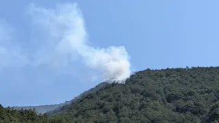 Ein Berg mit dichtem Wald stößt weißen Rauch aus, möglicherweise von einem Feuer. Der Himmel ist klar und blau.