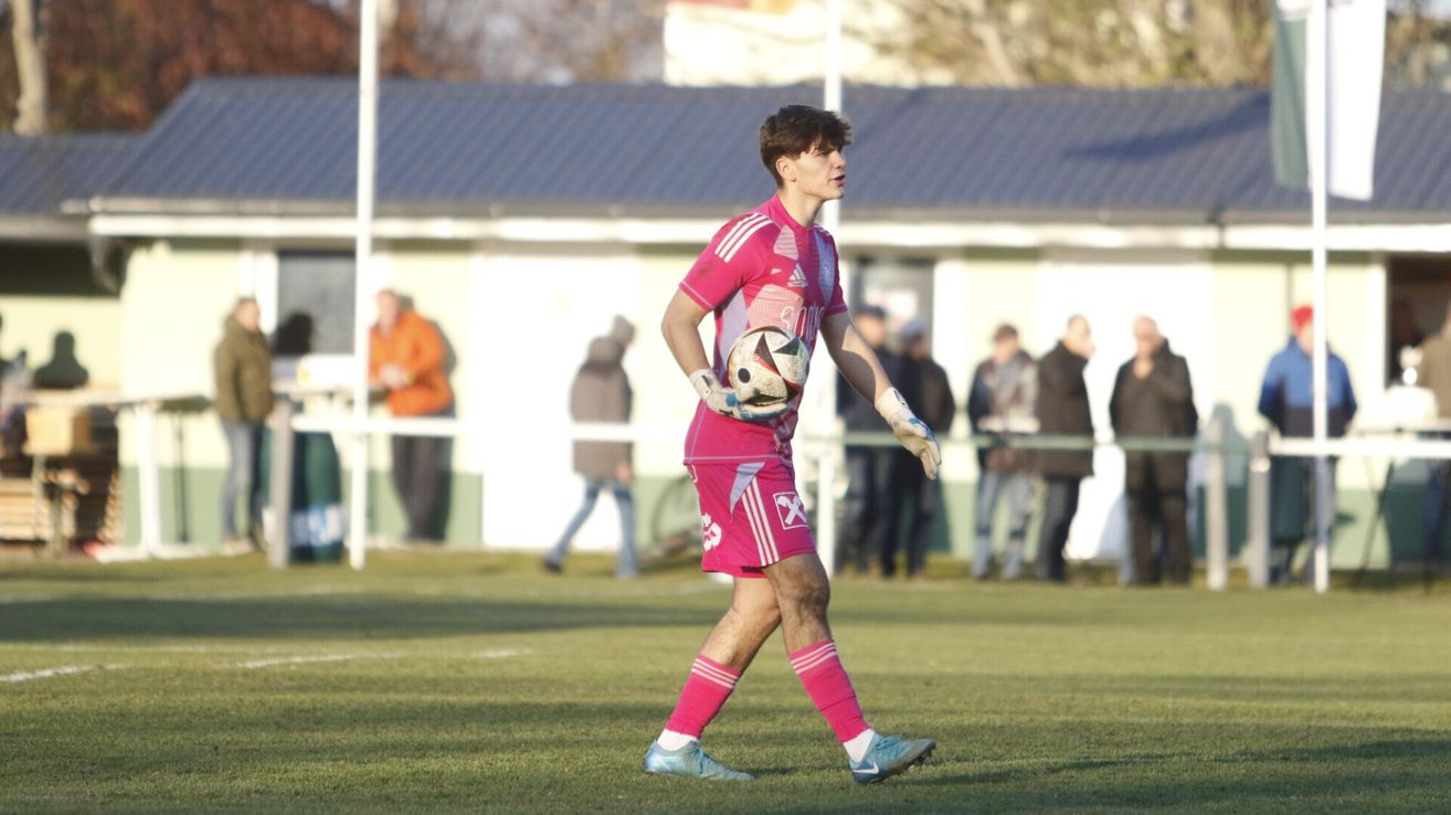 Ein Fußballspieler in einem rosa Trikot läuft auf einem Feld und hält einen Fußball. Dahinter stehen mehrere Personen und beobachten.