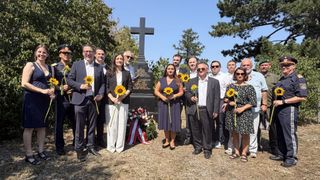 Eine Gruppe von Menschen steht vor einem Denkmal mit einem Kreuz, hält Sonnenblumen und ist feierlich gekleidet.
