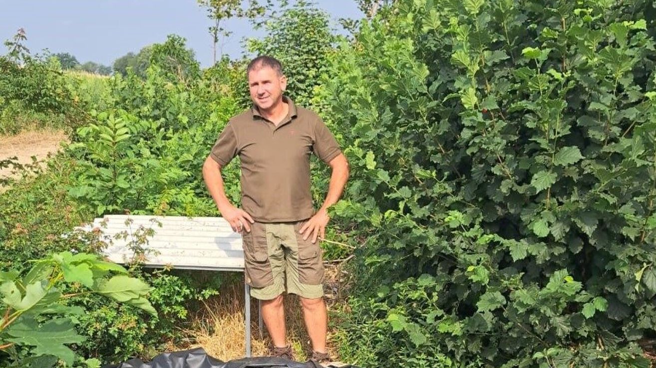 Ein Mann in einem braunen Hemd und Shorts steht in einem Garten mit üppiger Grünpflanze und einem weißen Tisch hinter ihm.