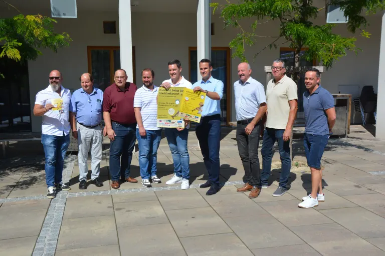 Eine Gruppe von Männern steht vor einem Gebäude, lächelt und posiert für ein Foto. Ein Mann hält ein gelbes Plakat.