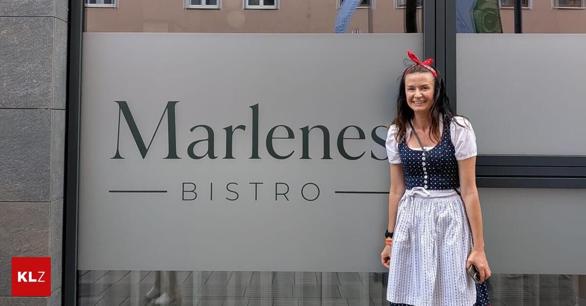 Eine Frau in traditioneller bayerischer Kleidung steht vor einem Schild des Marlenes Bistro und lächelt.