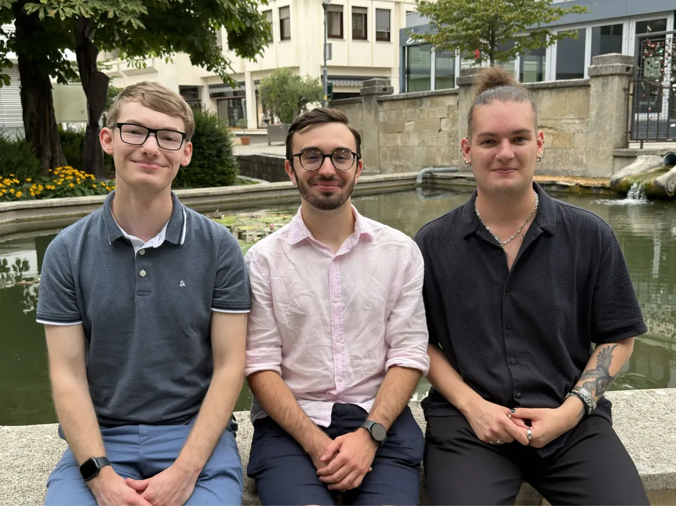Drei Männer sitzen auf einer Steinmauer in der Nähe eines Brunnens und lächeln für ein Foto. Der Mann auf der linken Seite trägt eine Brille und ein graues Poloshirt. Der Mann in der Mitte hat ein rosa Hemd und eine Uhr. Der Mann auf der rechten Seite trägt ein schwarzes Hemd und hat eine Halskette. Hinter ihnen steht ein Gebäude mit einem Baum.