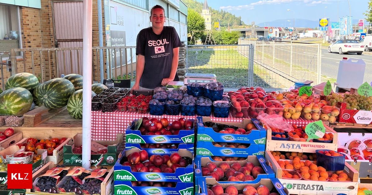 A man stands behind a table filled with various fruits, including peaches, plums, and watermelons, in a market setting.