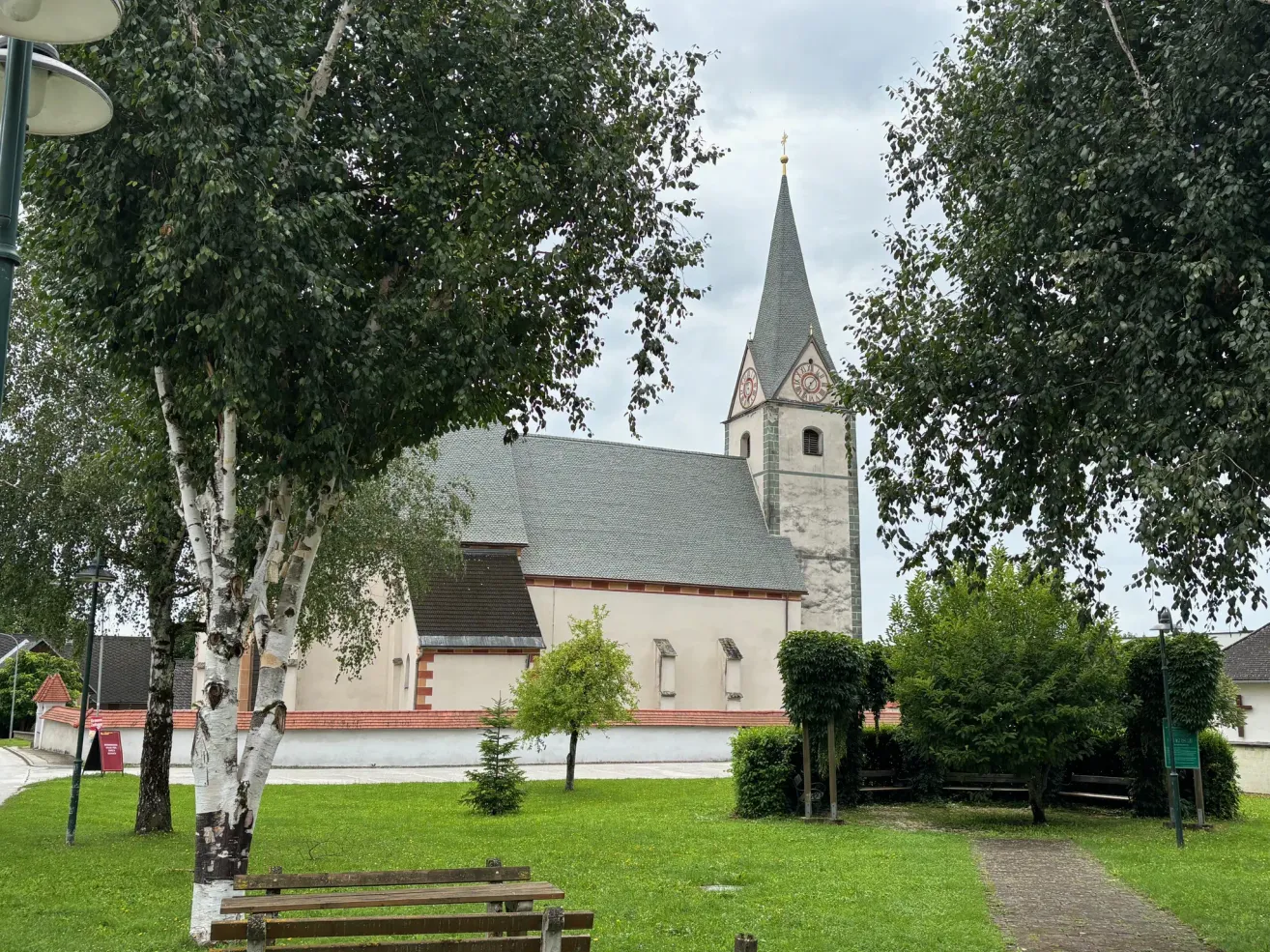 A small church with a steeple and clock faces, surrounded by lush greenery and trees.