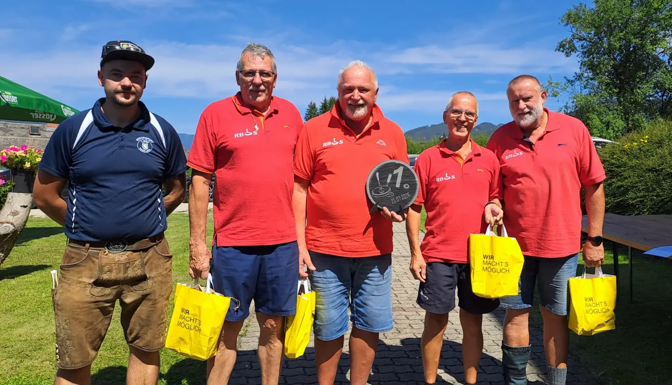 Fünf Männer in roten Hemden und blauen Shorts stehen lächelnd zusammen, einer hält eine Trophäe und andere halten gelbe Taschen. Sie sind draußen an einem sonnigen Tag mit Bergen im Hintergrund.