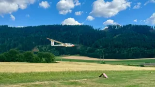 Ein Segelflugzeug mit der Nummer 12 fliegt über einem Grasfeld mit Bergen im Hintergrund.