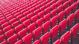 Ein Nahaufnahme eines Stadions mit Reihen roter Sitze, alle leer und ordentlich angeordnet. Die Sitze haben schwarze Armlehnen und sind in die gleiche Richtung ausgerichtet.