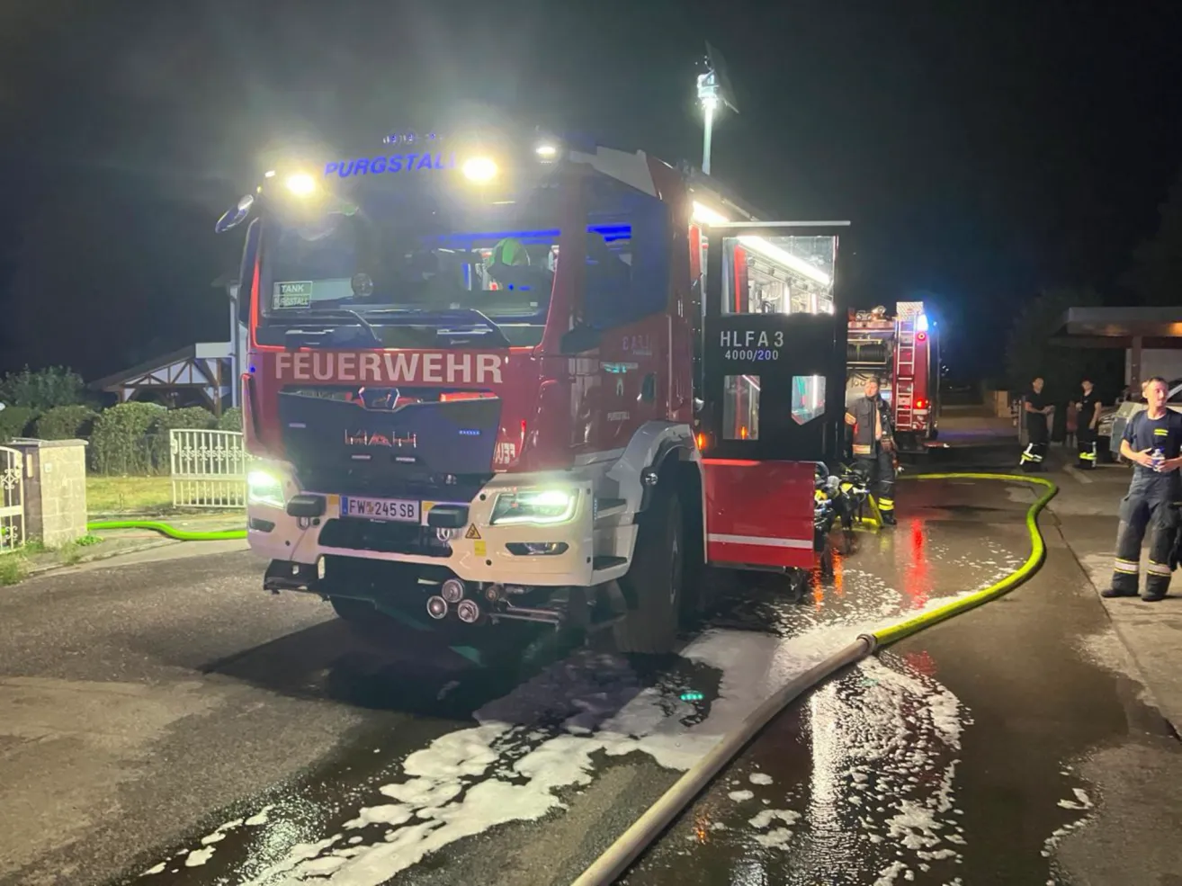 Ein roter Feuerwehrwagen mit dem Wort 'Feuerwehr' darauf ist am Straßenrand geparkt. Die Lichter sind eingeschaltet und der Wasserschlauch ist an den Wagen angeschlossen.