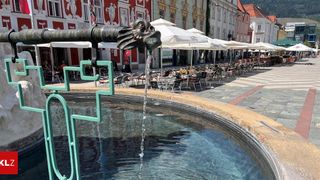 Ein Brunnen mit einem Drachenkopf, der Wasser in ein rundes Becken sprüht, umgeben von Tischen und Stühlen unter Schirmen auf einem Stadtplatz.