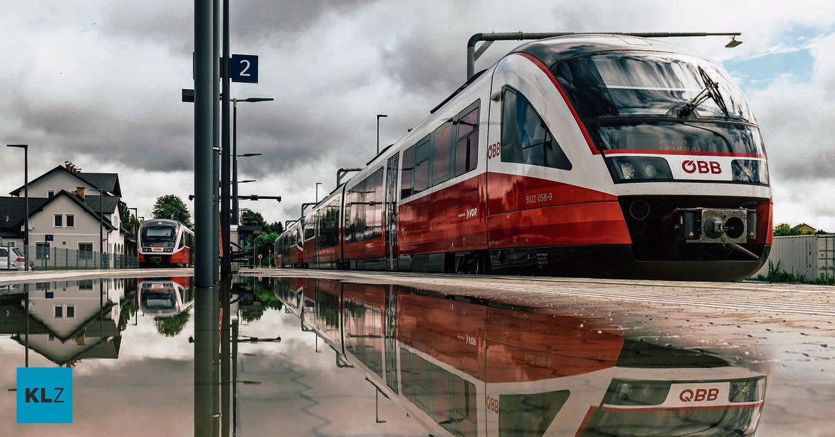 Ein roter und weißer Zug ist auf dem Bahnsteig eines Bahnhofs geparkt. Die Reflexion des Zuges ist auf dem nassen Bahnsteig sichtbar. Der Himmel ist bewölkt.