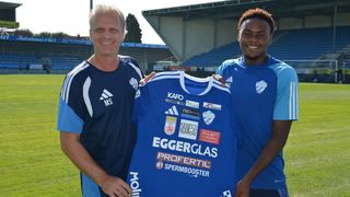 Drei Männer in blauen Fußballtrikots, einer hält ein Trikot mit dem Wort Eggerglas, stehen vor einem Stadion.